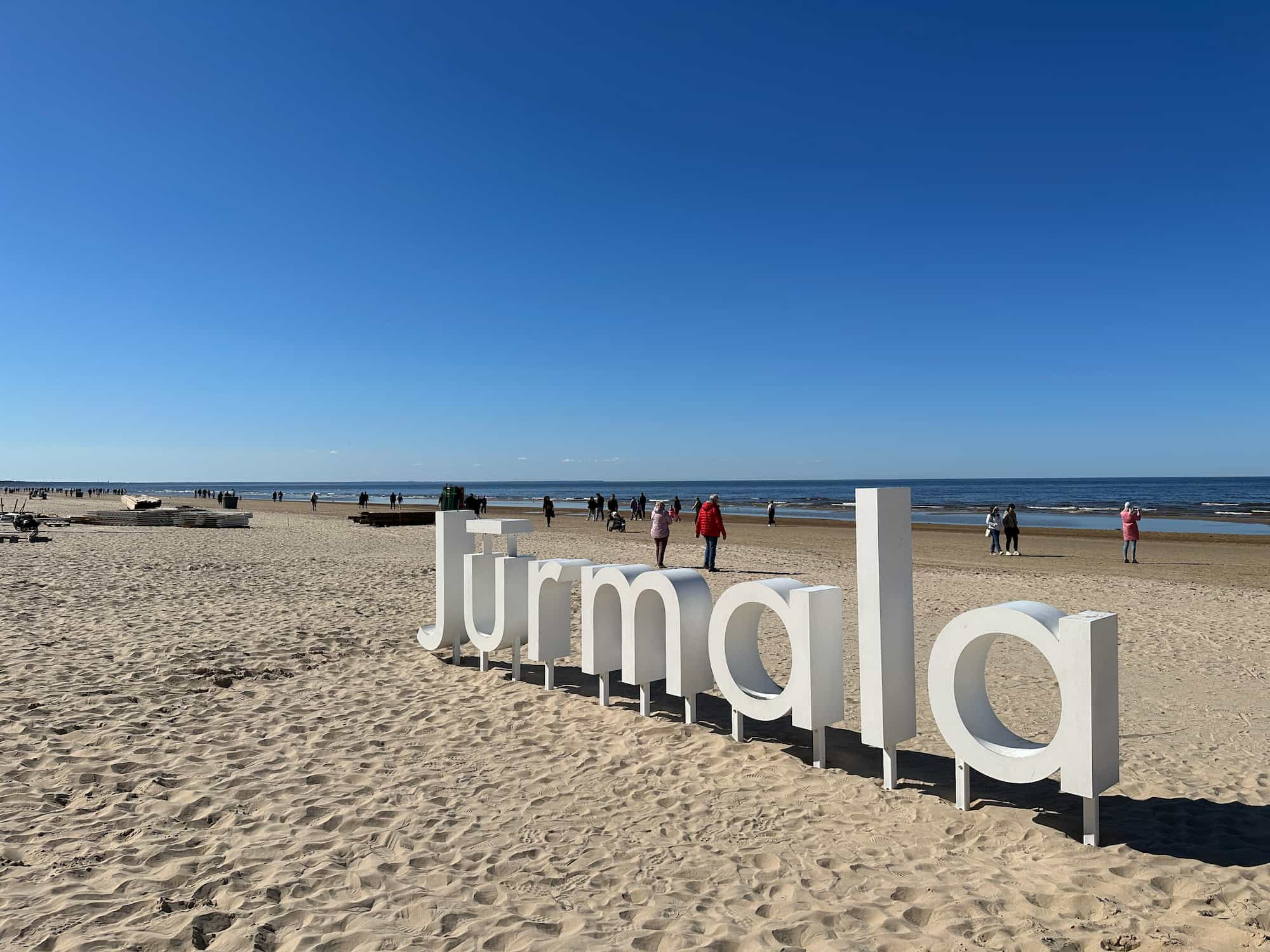 A big Hollywood-like sign on the beach. The sign says Jūrmala. People in warm jackets walking in the background